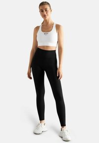 Valkoinen urheilu cropped-top logoilla, yhdistettynä korkeavyötäröisiin mustiin legginsseihin. Mallilla on päällään valkoiset lenkkarit. Sujuva kangas, tiukka malli.