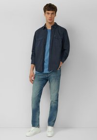 Dunkelblaues Hemd mit Knöpfen und zwei Brusttaschen über einem hellblauen T-Shirt, kombiniert mit hellblauen Jeans und weißen Sneakern. Einfaches, lässiges Design.