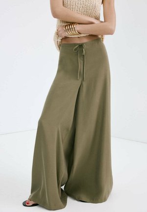 Luźne, oliwkowo-zielone spodnie szerokie w nogawkach z wiązaniem w talii, noszone z bezrękawnym beżowym swetrem i złotymi bransoletkami.