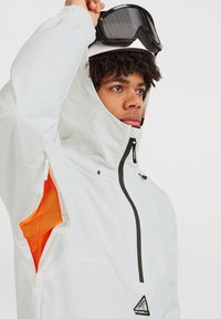 Witte sneeuwjas met een hoge kraag, zwarte rits en oranje mesh binnenvoering. Voorzien van een logopatch en een model dat skibril draagt.
