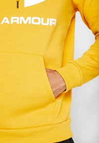 Sudadera amarilla con un bolsillo tipo canguro, puños y dobladillo acanalados. Presenta un gran logo blanco en el pecho. Tejido suave de mezcla de algodón.