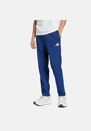 Mann trägt blaue Adidas-Trainingshose mit Logo am Oberschenkel, weiße Turnschuhe und ein weißes Hemd, steht mit einer Hand in der Tasche.