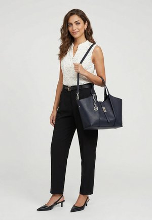 Vrouw draagt een mouwloze witte kanten top en zwarte broek, draagt een marineblauwe leren shopper met metalen hanger op de schouder.