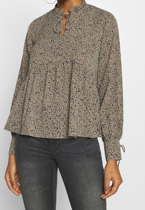 Locker sitzende beige Bluse mit navyfarbigem gesprenkeltem Muster, Rüschenausschnitt, Krawattendetail und gerafften Falten. Lange Ärmel mit elastischen Bündchen.