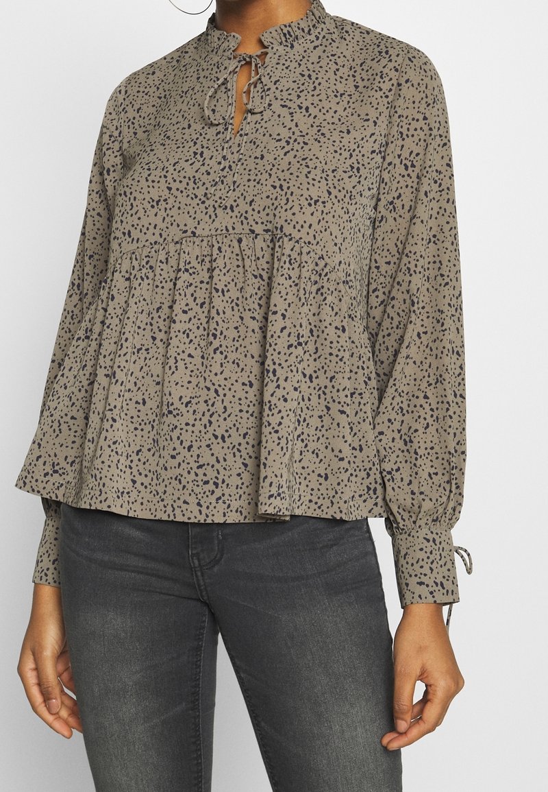 Blusa beige morbida con motivo a pois blu navy, scollatura arricciata, dettaglio con fiocco e pieghe arricciate. Maniche lunghe con polsini elasticizzati.