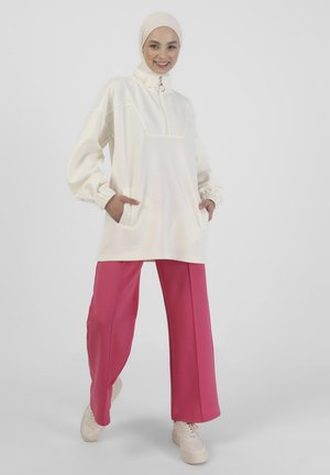 Suéter ligero color crema con cuello con cremallera, bolsillos frontales y puños elásticos. Combinado con pantalones de pierna ancha rosa y zapatos beige.