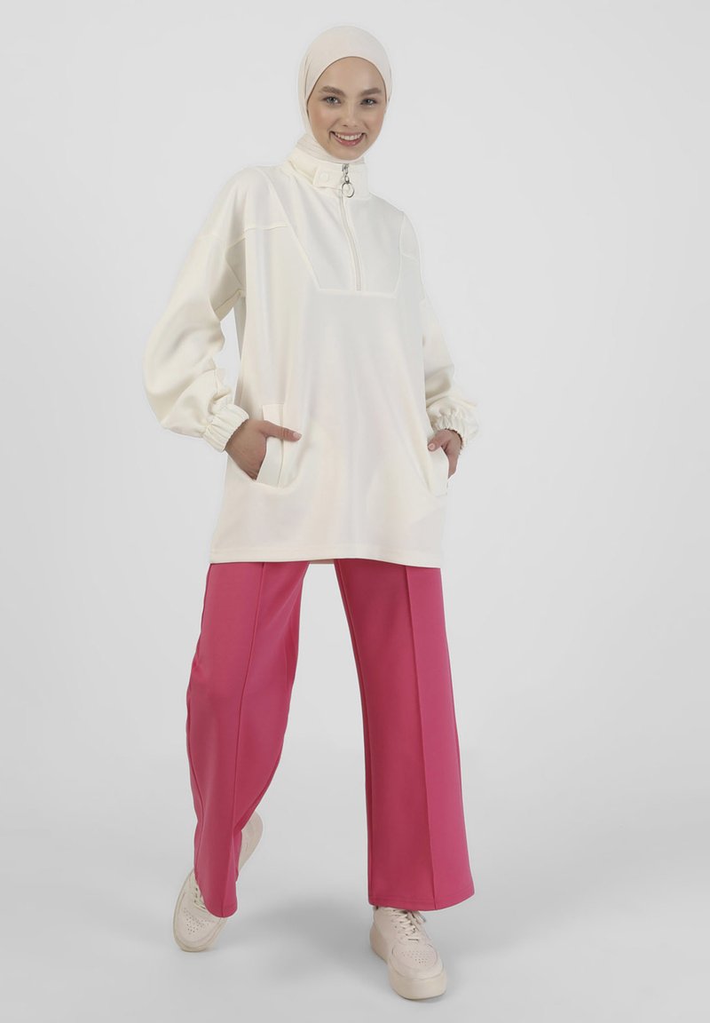 Pullover leggero di colore crema con collo con zip, tasche frontali e polsini elasticizzati. Abbinato a pantaloni larghi rosa e scarpe beige.