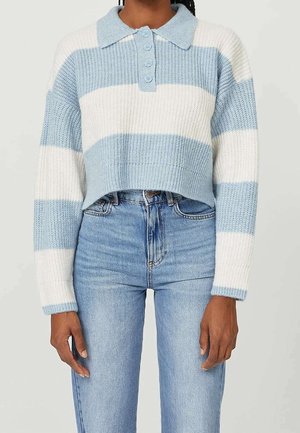 Femme portant un pull en maille rayé bleu clair et blanc, cropped, avec un col et des boutons, associé à un jean bleu clair taille haute.