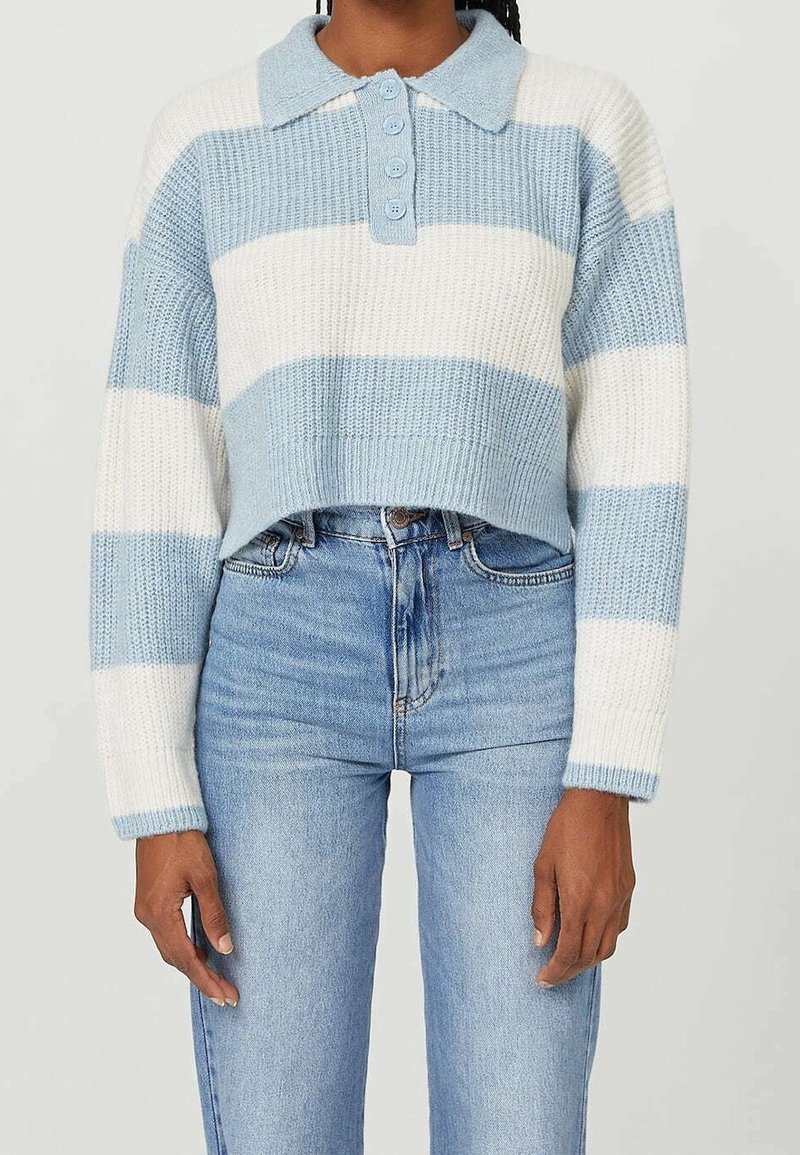 Donna che indossa un maglione a maglia strisciato blu chiaro e bianco, corto, con colletto e bottoni, abbinato a jeans blu chiaro a vita alta.