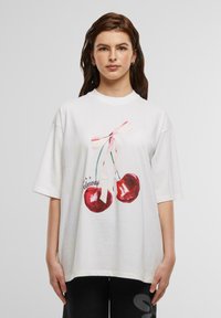 Hvit oversized T-skjorte med et grafisk trykk av to røde kirsebær med et rosa bånd og ordet "Cherious" i svart kursiv. Stoff i bomull.