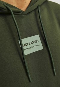 Sötétzöld kapucnis pulóver, elöl zsebbel és állítható húzózsinórokkal, egy téglalap alakú logófelirattal, amin a "JACK & JONES" felirat és egy szlogen található.