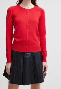 Woman wearing a fitted red buttoned cardigan and a black pleated leather skirt, standing against a plain light background.