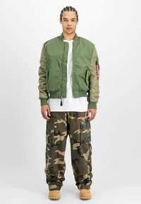 Bărbat cu părul împletit, purtând o jachetă bomber verde, un tricou alb, pantaloni cargo cu model camuflat și cizme bej, stând în fața unui fundal alb.