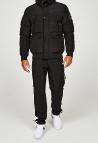 Sort dunjakke med høj krave, lynlås og trykknaplukning, flere lommer foran, båret sammen med sorte cargobukser og hvide sneakers.