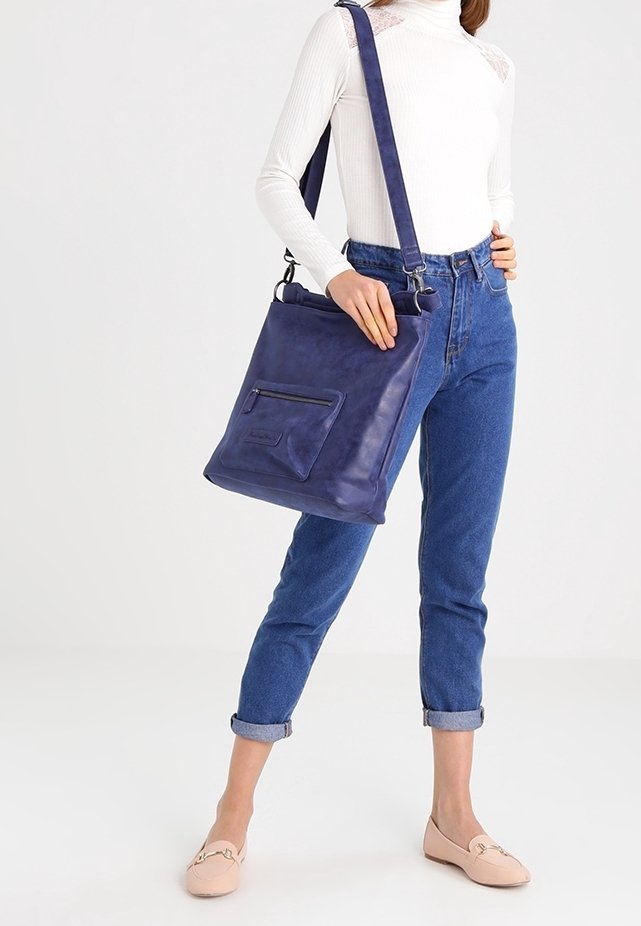 Femme portant un haut blanc à manches longues et un jean bleu tient un grand sac à bandoulière bleu marine avec une poche avant zippée, debout avec une main sur la hanche.