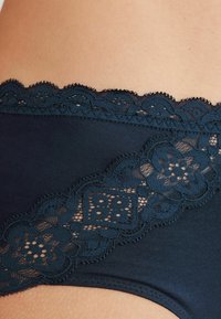 Donkerblauwe katoenen onderkleding met complexe kantdetails langs de tailleband met bloemenpatronen en golvende randen.