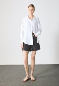 Hvid langærmet skjorte med knapper, parret med sorte denimshorts. Modellen har nude sandaler på. Enkel, minimalistisk design med en afslappet pasform.