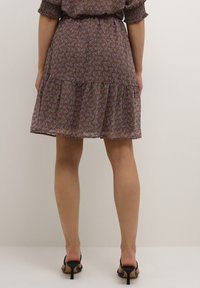 Žena v hnědých šatech ke kolenům s růžovým paisley vzorem a černých sandálech na podpatku s otevřenou špičkou, stojící na neutrálním pozadí.