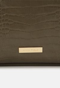 Olivgrüne, krokodilgeprägte Tasche aus synthetischem Material mit goldfarbener Markenplakette, glatt strukturiert und rechteckig geformt.