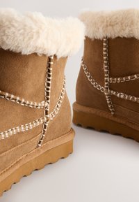 Bottes cheville en daim couleur fauve avec un revers en fausse fourrure crème duveteuse, des coutures décoratives, et une semelle en caoutchouc avec une adhérence texturée.