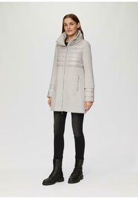 Helle graue Jacke mit gepolsterter Brustpartie, langen Ärmeln, hohem Kragen und seitlichen Taschen. Kombiniert mit schwarzen Skinny-Jeans und Schnürstiefeln.