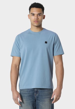 Ung mann iført en lys blå kortermet t-skjorte med en liten svart firkantet logo og falmede blå jeans, stående mot en hvit bakgrunn.