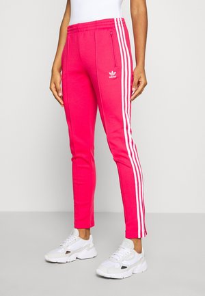 Persona che indossa pantaloni della tuta Adidas rosa acceso con strisce laterali bianche e scarpe da ginnastica bianche su uno sfondo chiaro e uniforme.
