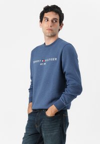 Mėlynas džemperis su apvalia apykakle, pagamintas iš medvilnės ir sintetinių pluoštų, turi baltą "TOMMY HILFIGER" logotipą ir tekstą "EST. 1985". Derinamas su tamsiomis džinsomis.