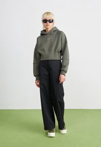 Joven vestido con gafas de sol negras, sudadera verde corta, pantalones anchos negros y zapatillas de plataforma verdes, de pie sobre una moqueta verde.