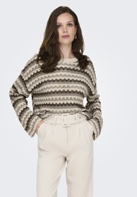 Strickpullover in Creme- und Brauntönen mit einem gewellten Muster. Lockere Passform, runder Ausschnitt und lange Ärmel. Kombiniert mit weißen, hoch taillierten Hosen.