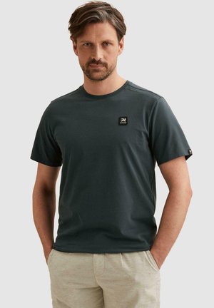 Man met kort bruin haar en baard, draagt een donker groen T-shirt en licht beige broek, handen in de zakken, neutrale achtergrond.