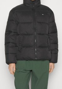 Svart pufferjacka med hög krage, kviltat mönster, dragkedja fram och sidofickor. Bärs över gröna texturerade byxor.