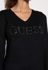 Schwarzer Pullover mit V-Ausschnitt, auf dem "GUESS" mit Strasssteinen auf der Vorderseite verziert ist, und einer glatten Textur.