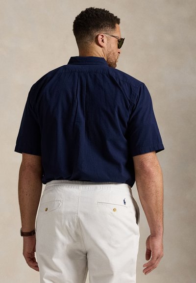 Homme avec des cheveux courts et bouclés portant des lunettes de soleil, une chemise bleu marine et un pantalon blanc, de dos contre un fond neutre.