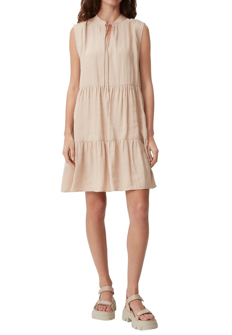 Robe sans manches en mélange de coton beige clair avec un col en forme d'œillet, un bas à volants et une silhouette fluide, portée avec des sandales épaisses.