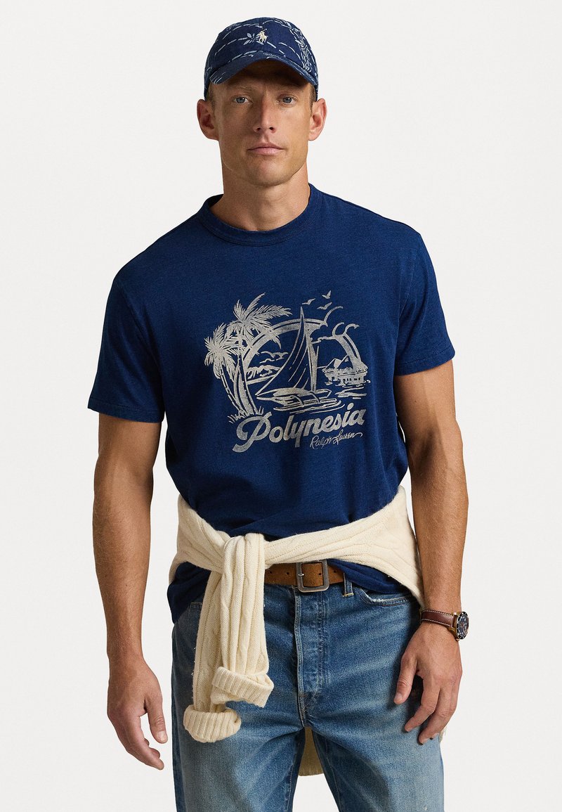 Mörkblå t-shirt med grafik av en palm och segelbåt i ljusgrått. Modellen bär en krämfärgad tröja knuten i midjan och denimjeans.