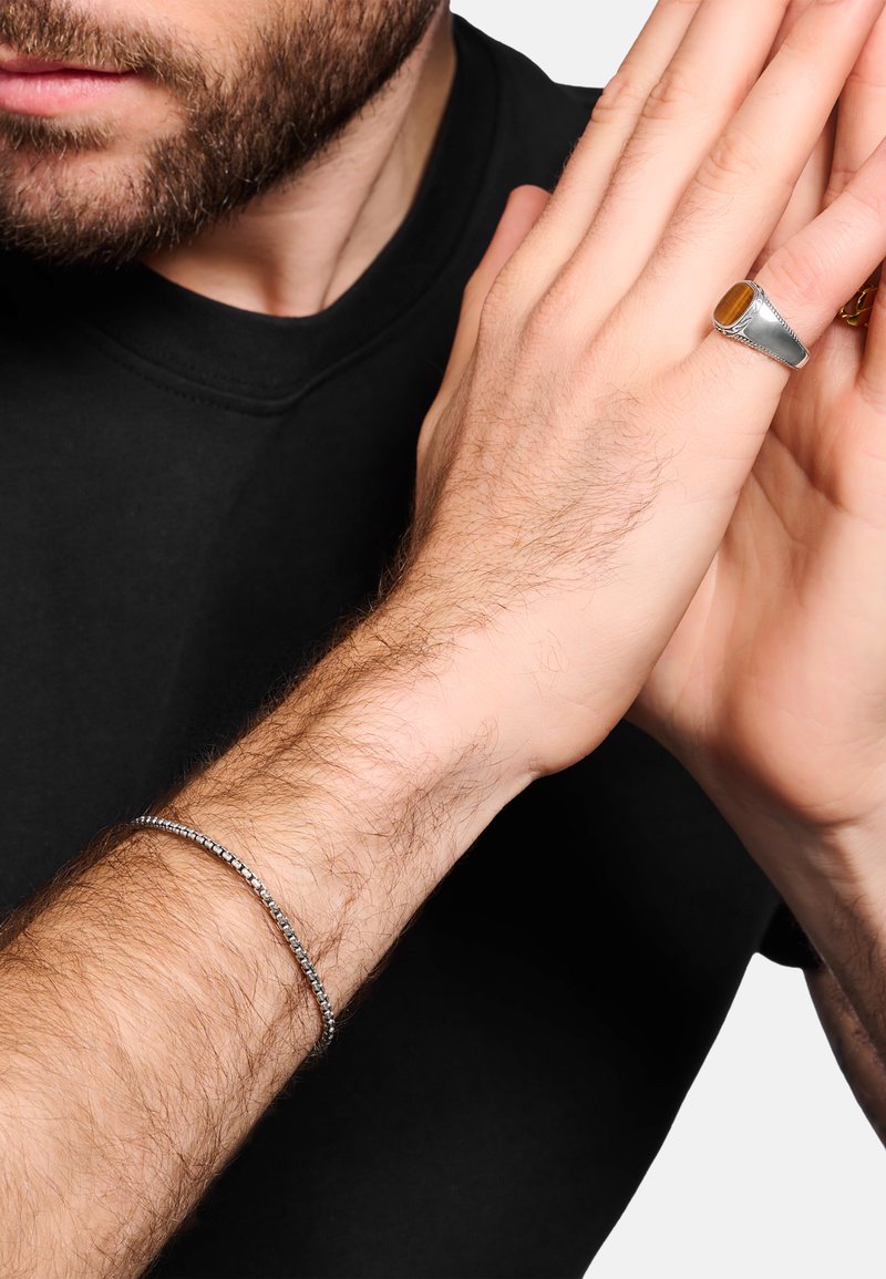 Silberarmband am Handgelenk, glatt mit kleinen runden Akzenten. Trägt einen Silberring mit einem gelben Stein an einer Hand, die nach vorne positioniert ist.