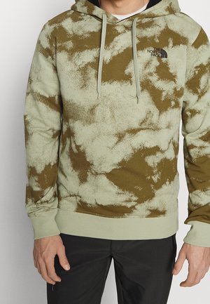 Uomo con felpa con cappuccio tie-dye verde e marrone con il logo The North Face, pantaloni neri, in piedi davanti a uno sfondo semplice.