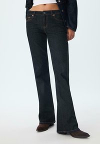 Jeans en denim foncé évasés avec des coutures orange, dotés d'une fermeture à bouton et de poches avant, associés à des bottes à talons marron.