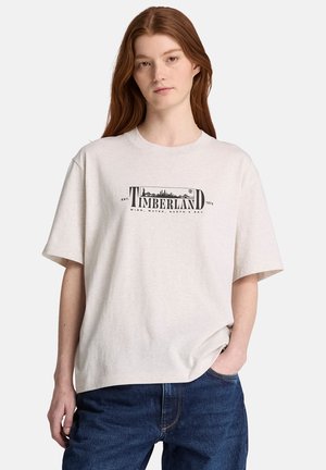Ung kvinde med langt rødt hår iført en lysebeige Timberland T-shirt og blå jeans, den ene hånd i lommen, neutral udtryk.
