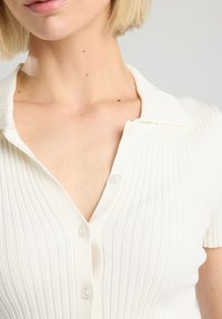 White ribbed polo shirt featuring a v-neckline, short sleeves, and button detailing along the front. Soft texture with vertical rib pattern.