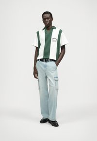 Groen en wit gestreepte polo met korte mouwen, v-hals en logo; gecombineerd met lichtblauwe cargobroek en zwarte schoenen.