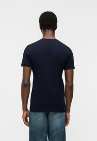 Άνδρας με κοντά σγουρά μαλλιά, που φοράει απλό σκούρο μπλε μπλουζάκι και μ πλε τζιν, στέκεται με την πλάτη στραμμένη σε λευκό φόντο.