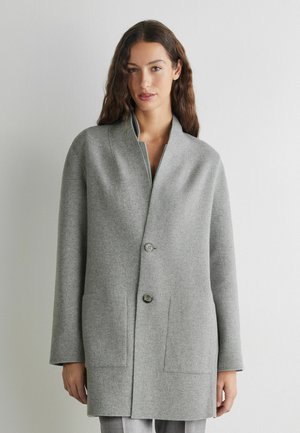 Femme aux longs cheveux bruns, portant un manteau en laine gris clair boutonné avec des poches plaquées, debout devant un fond blanc uni.