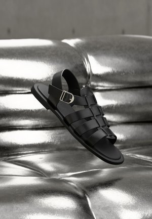 Schwarze Lederfisherman-Sandale mit silberner Schnalle, die über einer metallisch-silbernen, gepolsterten Oberfläche schwebt.