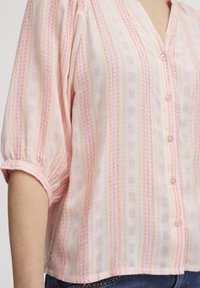 Blusa rosa chiaro con strisce verticali in tonalità più scure, tessuto strutturato con un motivo intessuto, chiusura a bottoni e maniche corte a sbuffo.