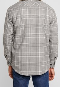 Rutigt skjorta med grå och vit schackmönster, svarta accenter, knäppta manschetter och en mjuk krage. Tyget ser texturerat ut.