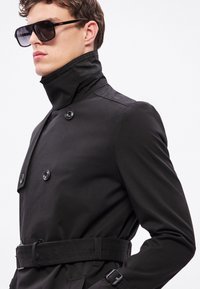 Must kahepoolse nööpimisega trentšmantel kõrge kraega, millel on suured nupud ja sobiv vöö. Modellil on tumedad päikeseprillid.