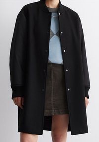 Cappotto lungo nero con colletto rotondo, bottoni a scatto e polsini a coste; indossato sopra un maglione blu con motivo e una gonna in velluto a coste.