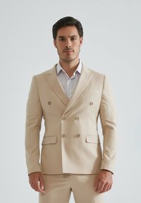 Veste de costume beige à double boutonnage en tissu lisse, dotée de deux poches avant, de boutons assortis et d'une silhouette ajustée.